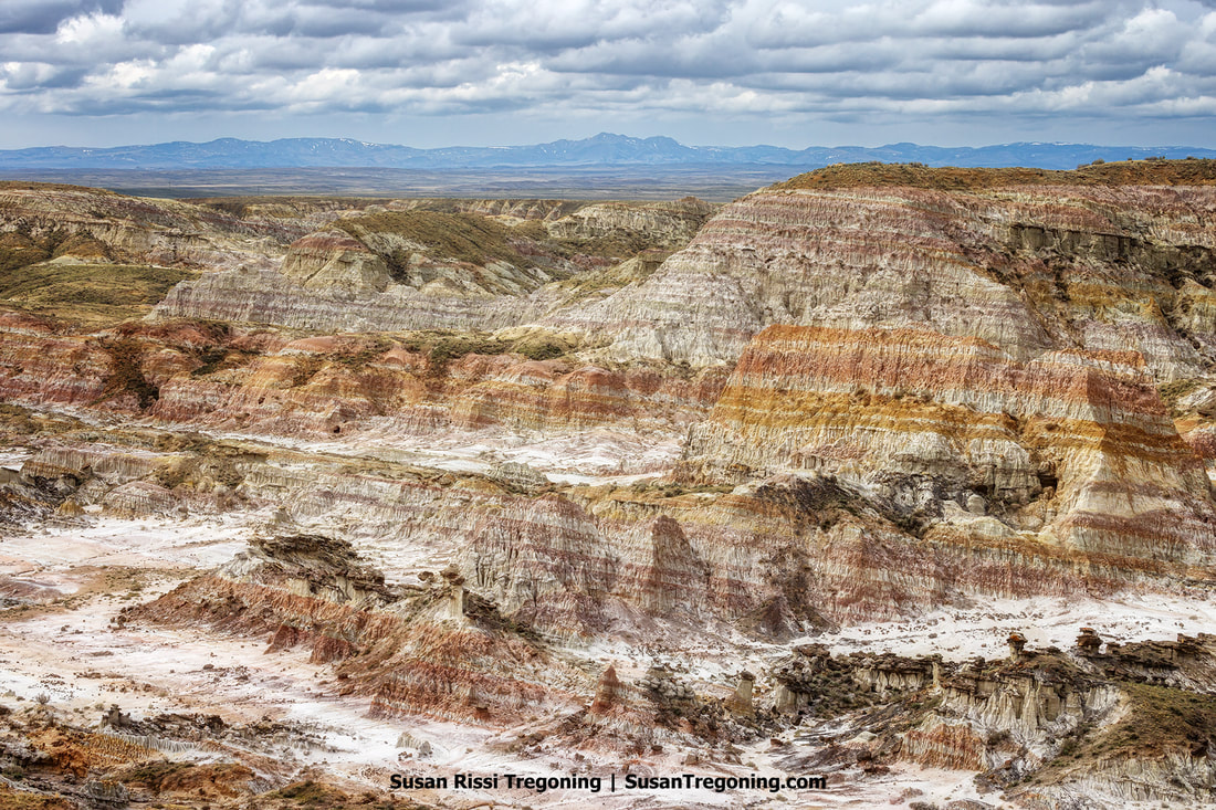 In expansive badlands, variegated layers of earth reveal millions of years of geological history. Overlooking the rugged terrain, the cloudy sky adds a dramatic backdrop to the striking natural features of Wyoming's otherworldly Hells Half Acre landscape.